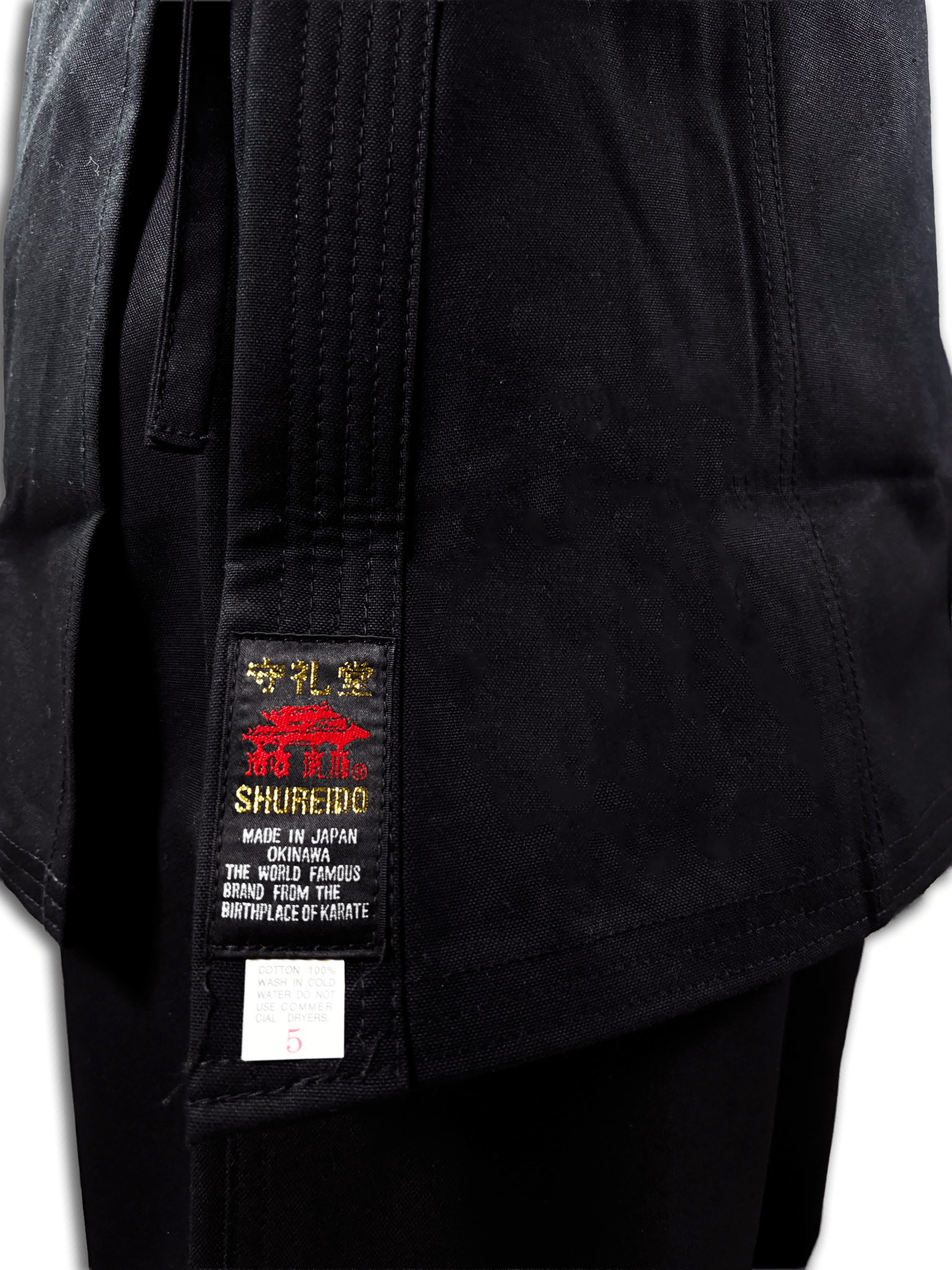 Detailed close-up of the Shureido Okinawa brand label and 100% cotton tag on the hem of a black karate gi jacket and belt.