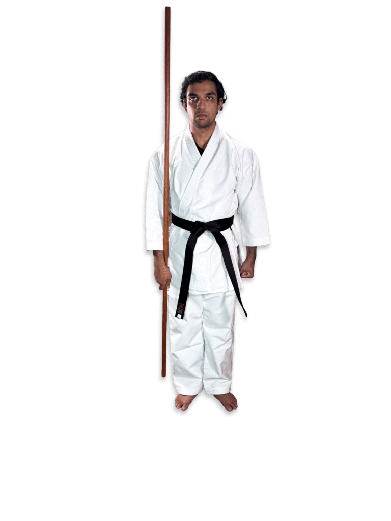 Martial artist in white karate gi and black belt holding a traditional Shureido Okinawan wooden bo staff in a ready stance against a clean background.