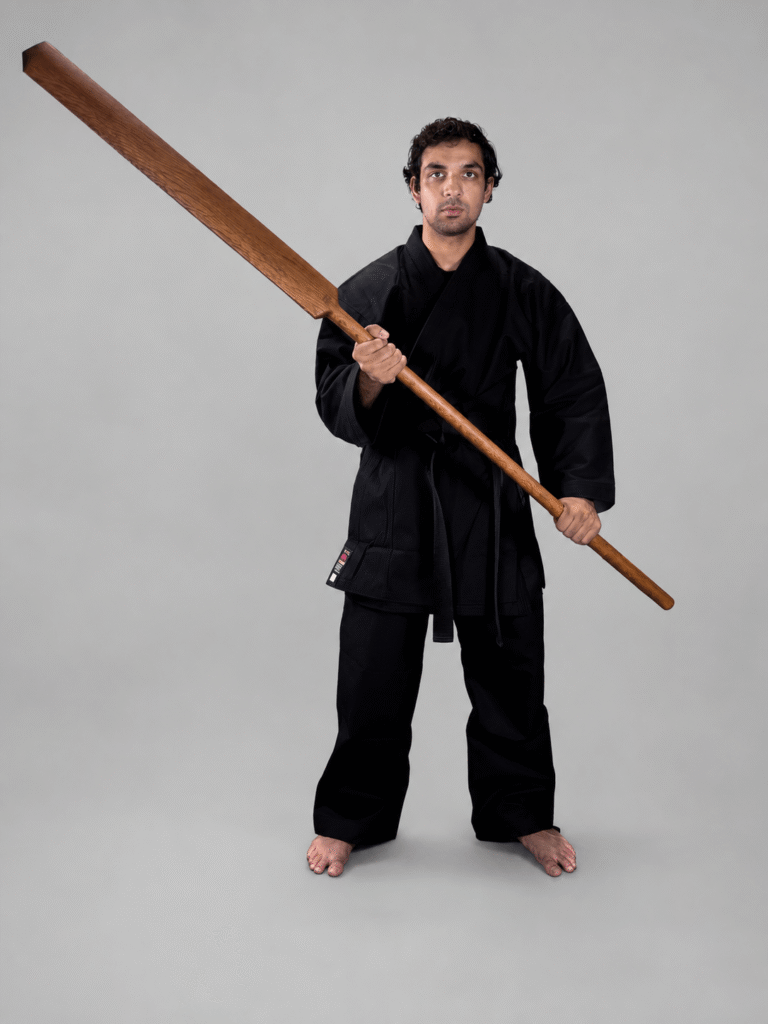 Martial artist wearing a black karate gi and black belt standing upright and holding a traditional Shureido Okinawan wooden eku oar weapon