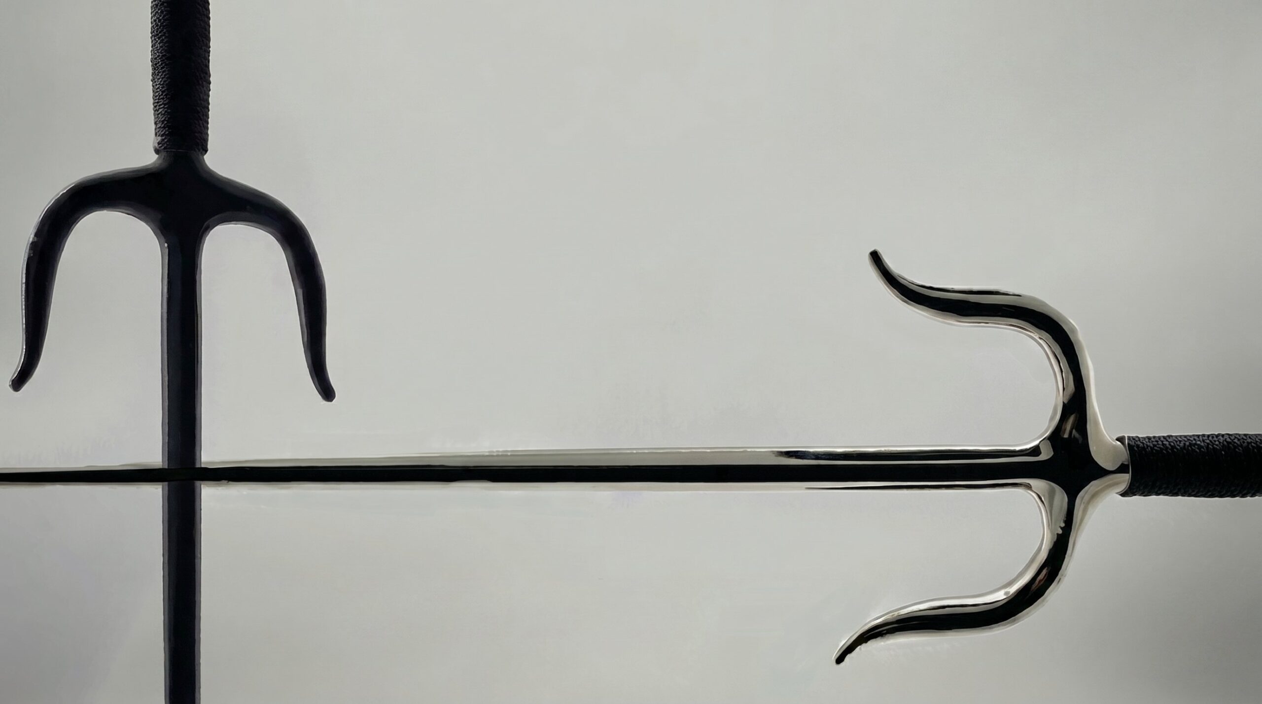 Two traditional Okinawan sai weapons with black wrapped handles crossed against a clean, light grey background.